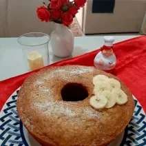 Fotografia em tons de azul em uma mesa com um pano vermelho, um prato redondo raso com desenho em azul e um bolo de banana com canela e rodelas de banana em cima dele.