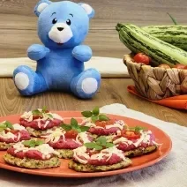 Fotografia de uma mesa de madeira com um prato laranja apoiado em um paninho branco. Sobre esse prato tem sete unidades de mini esfihas abertas com molho de beterraba, queijo, tomate e manjericão. No fundo tem o ursinho azul de mucilon e vegetais.