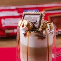 Fotografia em tons de vermelho com uma caneca de vidro ao centro. Dentro da caneca existe uma receita de café com leite finalizada com marshmallow e Choco Biscuit NESTLÉ