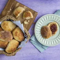 Fotografia em tons de roxo e azul em uma bancada de madeira roxa com uma tábua de madeira e uma cesta de vime com vários pãeszinhos dentro dele e um cortado ao meio ao lado da tábua. Ao lado, um pano azul claro com um prato azul e uma fatia de pão.