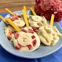 Fotografia em tons de azul e rosa com um prato azul ao centro. Em cima do prato existe vários picolés feitos de iogurte e frutas.