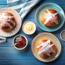 Fotografia vista de cima de três pratos com cor diferente, e em ada um, tem um pão doce recheado com coco e cobertura brilhante de açúcar e glacê. Ao lado tem alguns recipientes pequenos de vidro brancos com confeito, creme branco e no outro, geleia