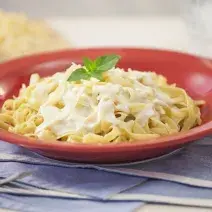 Fotografia em tons de vermelho e azul em uma bancada de madeira com jogos americanos azul e um prato vermelho com o macarrão ao molho branco dentro. Ao fundo, uma peça de queijo parmesão em fatias.