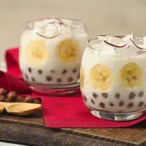 Fotografia em tons de vermelho e marrom em uma bancada branca com uma tábua de madeira escura, um pano vermelho, duas taças de vidro com o docinho de banana com Nescau Cereal.