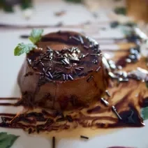 Fotografia de um mini pudim de chocolate sem furo no meio, decorado com granulado, calda de chocolate e uma folha de hortelã, sobre um prato branco desenhado..