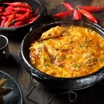 Fotografia de uma panela de barro com caldeirada borbulhando de peixe com cação, postas de pescada e corvina, molho de tomate, açafrão, louro e salsinha.