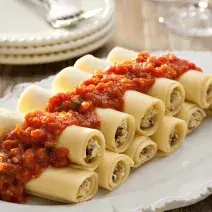 Fotografia em tons de vermelho em uma bancada de madeira com um recipiente oval branco grande com vários canelones de berinjela e com molho de tomate em cima dele.