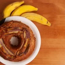 Fotografia em tons de marrom claro com um prato branco ao centro. Dentro do prato existe um bolo de banana redondo com um furo no meio, por cima do bolo existe pedaços de bananas caramelizadas. Ao lado existe três bananas inteiras.