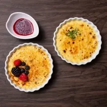 Fotografia vista de cima de dois cremes brulée, um de alho poró com queijo canastra e o outro, sabor cumaru com pistache e frutas vermelhas, os dois cremes brulée estão em ramekins. Ao lado tem uma calda de frutas vermelhas.