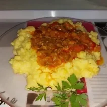 Fotografia de um prato raso transparente com uma polenta caseira. Por cima tem molho de tomate e, ao lado, tem salsinha.