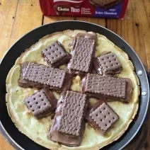 Fotografia em tons de marrom e vermelho com um prato preto ao centro. Em cima do prato tem uma pizza feita de massa de ovo e tapioca. Por cima da massa existe vários biscoitos Choco Trio derretidos.