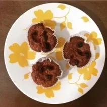 Fotografia vista de cima de três forminhas brancas com bolinho de banana e damascos por cima. As forminhas estão sobre um prato branco com flores amarelas.