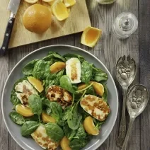 Fotografia vista de cima de uma salada de agrião, frango defumado e gomos de laranja, que está em um prato fundo cinza, sobre uma mesa de madeira. Ao lado, uma tábua de madeira de tom claro, com uma laranja inteira, cascas da fruta e uma faca apoiada.