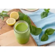 Fotografia de um copo de vidro decorativo com suco detox de cor verde feito com couve, maçã verde, pepino, gengibre e nutren. O copo está sobre uma tábua de madeira, ao lado de folhas de hortelã e metades de limão siciliano sobre a mesa.