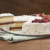Fotografia em tons de marrom, verde e branco de uma bancada de madeira escura com um paninho verde, sobre ele um prato branco redondo com um bolo recheado e coberto com frutas vermelhas.