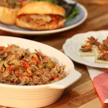 fotografia tirada de uma travessa branca com carne desfiada dentro com pedaços de azeitonas ao lado um prato redondo e branco com a carne desfiada e pedaços de azeitonas e abaixo um pano branco e vermelho