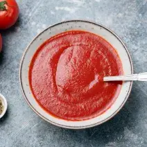 fotografia em tons de cinza e vermelho de uma bancada cinza vista de cima. Ao centro um recipiente redondo com sopa de tomate e um colher para servir, e ao lado esquerdo contém tomates.