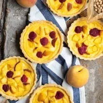 Fotografia vista de cima de cinco minitortinhas com um creme de frutas, e pedaços por cima, sobre uma toalha de mesa na cor branco e azul. Ao redor, alguns pêssegos e grãos de bico dentro de um saquinho de papel manteiga.