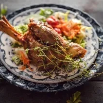 Fotografia de um cordeiro temperado e assado com vinho e ervas e um prato azul e branco, e embaixo da carne, uma salada colorida. Ao redor do prato, algumas folhas de ervas sobre uma mesa de madeira em tom escuro.