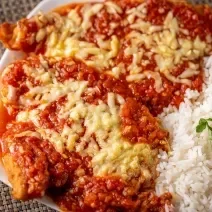 Fotografia de um prato retangular branco com dois filés de frango à parmegiana com queijo derretido por cima, ao lado de um pouco de arroz e batatas fritas.