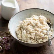 Fotografia em tons claros de um recipiente de vidro branco com acaçá cremoso, e no mesmo recipiente, uma colher de sopa apoiada dentro do acaçá. Ao fundo, molho em um recipiente pequeno na cor branca, sobre uma mesa de madeira na cor marrom escuro.