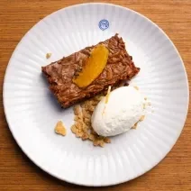 Fotografia de um prato branco raso com um pedaço de bolo de chocolate cremoso com crocante de nozes, e por cima, chantilly de laranja. O prato está sobre uma bancada de madeira clara.