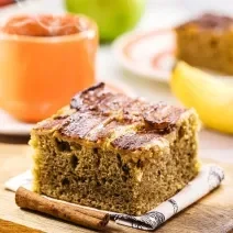 Fotografia de um pedaço de bolo de banana saudável, apoiado em um paninho branco, ao lado de dois paus de canela, sobre uma tábua pequena de madeira. Ao fundo tem uma caneca laranja e uma banana.