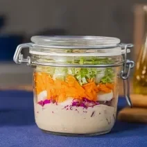 Fotografia em tons de azul com um pote de vidro ao centro. Dentro do pote existe uma receita de salada de repolho roxo feita com repolho, cenoura, palmito e molho rosé