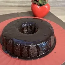 Fotografia de um bolo de chocolate e café com calda de chocolate sobre um apoio grande vermelho. O apoio está por cima de um pano marrom de tom escuro.