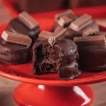 Fotografia em tons de vermelho em uma bancada de madeira e um prato vermelho redondo ao centro. Em cima dele, existem pães de mel de chocolate com KitKat em cima decorando e um aberto mostrando o recheio.