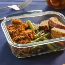 A fotografia mostra uma marmita de vidro com carne desfiada dourada, batata-doce assada levemente crocante e vagem refogada vibrante, organizada de forma prática e equilibrada, com cores intensas e aparência convidativa.