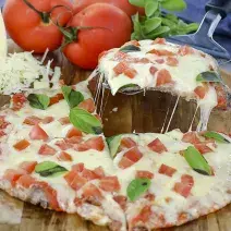 Foto aproximada de uma pizza marguerita, decorada com tomates e folhas de manjericão, sobre uma tábua de madeira e com tomates ao fundo