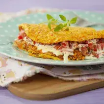 Fotografia em tons de roxo e azul em uma mesa roxa e uma tábua de madeira com um paninho de ursinhos coloridos apoiado nela. Em cima, um prato azul de bolinhas brancas com a crepioca de cenoura com recheio de tomate e requeijão.