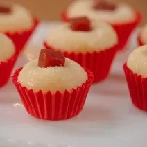 Fotografia de sete unidades de forminhas vermelhas com brigadeiro de romeu e julieta. Por cima de cada um tem um quadradinho de goiabada, e todos estão sobre um apoio decorativo de cor branca.