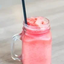 Fotografia de um refresco de melancia com leite MOÇA e gelo, está em um copo de vidro decorativo com alça e um canudo preto dentro.