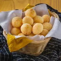 Foto aproximada de uma cesta de vime com diversos pães de queijo dentro. Ela está sobre uma bancada de madeira com um tecido azul quadriculado.