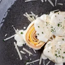 Fotografia vista de cima de três unidades de rondelli deitados em um prato raso escuro. Por cima das massas tem molho branco com queijo parmesão ralado. O prato está sobre uma mesa de madeira preta.