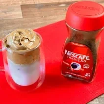 Foto aproximada da receita de Dalgona Café feito com Nescafé Tradição, servido em um xícara grande transparente ao lado de um pote de Nescafé, tudo sobre um pano vermelho em uma tábua de madeira