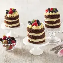 Fotografia em tons de branco e cinza de uma bancada cinza com com suportes de bolos brancos e de vidro com naked cakes. Ao lado um paninho rosa com garfos e um recipiente redondo de vidro com frutas vermelhas.