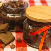 Foto de uma bancada forrada com uma toalha quadriculada vermelha e branca. Sobre ela há 2 potes de vidros com a receita de Brownie de Nescau com Brigadeiro. Um deles está fechado e o outro aberto. Além disso há 2 biscoitos e algumas bolinhas de cereal