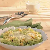 Fotografia em tons de verde em uma bancada de madeira clara, um pano verde, um pegador de salada e um prato redondo branco fundo com a salada de alface, queijo, torradinhas e iogurte dentro.