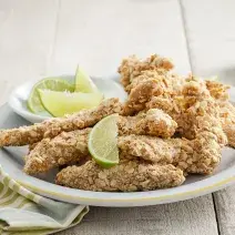 Fotografia em tons de verde em uma bancada de madeira clara, um pano com listras verdes, um prato branco raso com vários filés de frango empanados e fatias de limão em cima dele.