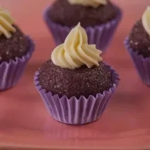 Fotografia de quatro forminhas de cor roxa com um brigadeiro de açaí, e por cima de cada um tem uma cobertura de chocolate confeitado. As unidades de brigadeiro estão sobre um apoio de cor rosa.