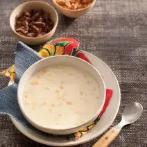 Fotografia em tons de cinza e  branco de um paninho cinza, sobre ele um prato redondo branco raso  com um paninho estampado, sobre ele um prato redondo fundo branco com a canjica. Ao lado uma faca com cabo de madeira. Ao fundo dois potinhos com