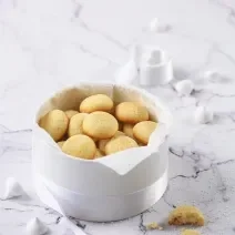 Fotografia em tons de branco de alguns biscoitos de massa branca dentro de uma cestinha branca com uma fita também branca amarrando-a. Ao fundo, alguns suspiros e um biscoito quebrado ao meio, sobre uma mesa de mármore branca.