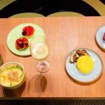 Fotografia de uma mesa de madeira com um brunch com cuscuz, quiche de alho poró, cheesecake, salada de frutas e suco de abacaxi com hortelã. As comidas estão em pratos coloridos, e o suco estão em taças, na imagem são três.