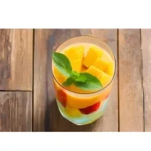 Fotografia de um copo de vidro com um refresco de frutas, com pedaços de frutas e por cima, folhas de hortelã. O copo está sobre uma mesa de madeira.