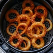 Fotografia vista de cima de várias onion rings dentro da cesta da air fryer, que está sobre uma mesa de madeira.