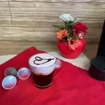 Fotografia em tons de vermelho em uma bancada de madeira, um pano vermelho e uma xícara com café e espuma rosa decorado com um coração com calda de chocolate ao centro. Ao fundo, um vaso vermelho com flores e em formato de coração.