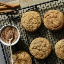 Fotografia vista de cima de sete cookies com açúcar e canela sobre uma grade preta. Ao lado dos cookies e por cima da grade, tem um pequeno pote de vidro branco com açúcar e canela, e uma colher de chá apoiada sobre ele.